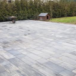 ABERBRUK - Nowo ułożona, wilgotna nawierzchnia z szarych prostokątnych płyt betonowych, z ceglanymi słupkami ogrodzenia i taczką w tle oraz budą dla psa, otoczona zielenią.