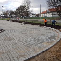 ABERBRUK - Układanie szarej kostki brukowej na parkingu sklepu, widoczni pracownicy w odblaskowych ubraniach wyrównujący ziemię pod nasadzenia wzdłuż obrzeża.