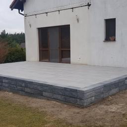 ABERBRUK - Szary taras z kostki brukowej, otoczony obrzeżem z ciemniejszej kostki, z niskimi schodami, przylegający do białego budynku z brązowymi oknami.