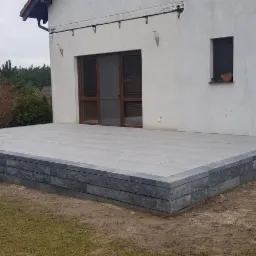 Szary taras z kostki brukowej, otoczony obrzeżem z ciemniejszej kostki, z niskimi schodami, przylegający do białego budynku z brązowymi oknami.