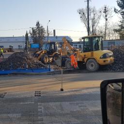 ABERBRUK - Układanie szarej kostki brukowej na placu przed budynkiem, z widocznymi maszynami budowlanymi i stertą gruzu, pracownicy w odblaskowych kamizelkach w tle, częściowo widoczne odbicie pracownika...