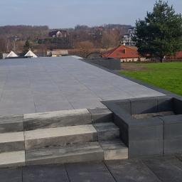 ABERBRUK - Nowoczesny taras z jasnoszarych płyt, schody z kostki o melanżowym wzorze szarości, donice z ciemnej kostki betonowej, w tle widok na zalesione wzgórza i zabudowania.