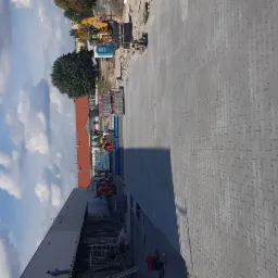 Układanie szarej kostki betonowej typu 'I' na dużej powierzchni placu budowy w słoneczny dzień, z widocznym ciężkim sprzętem i częściowo ukończonym budynkiem w tle.