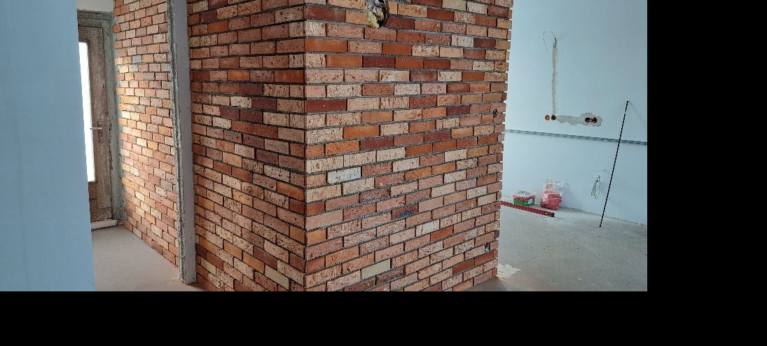 Ściana wykończona czerwoną cegłą klinkierową w rogu pomieszczenia. Widoczne przewody elektryczne w ścianie i poziomica na podłodze. Wykończenie w trakcie realizacji.
