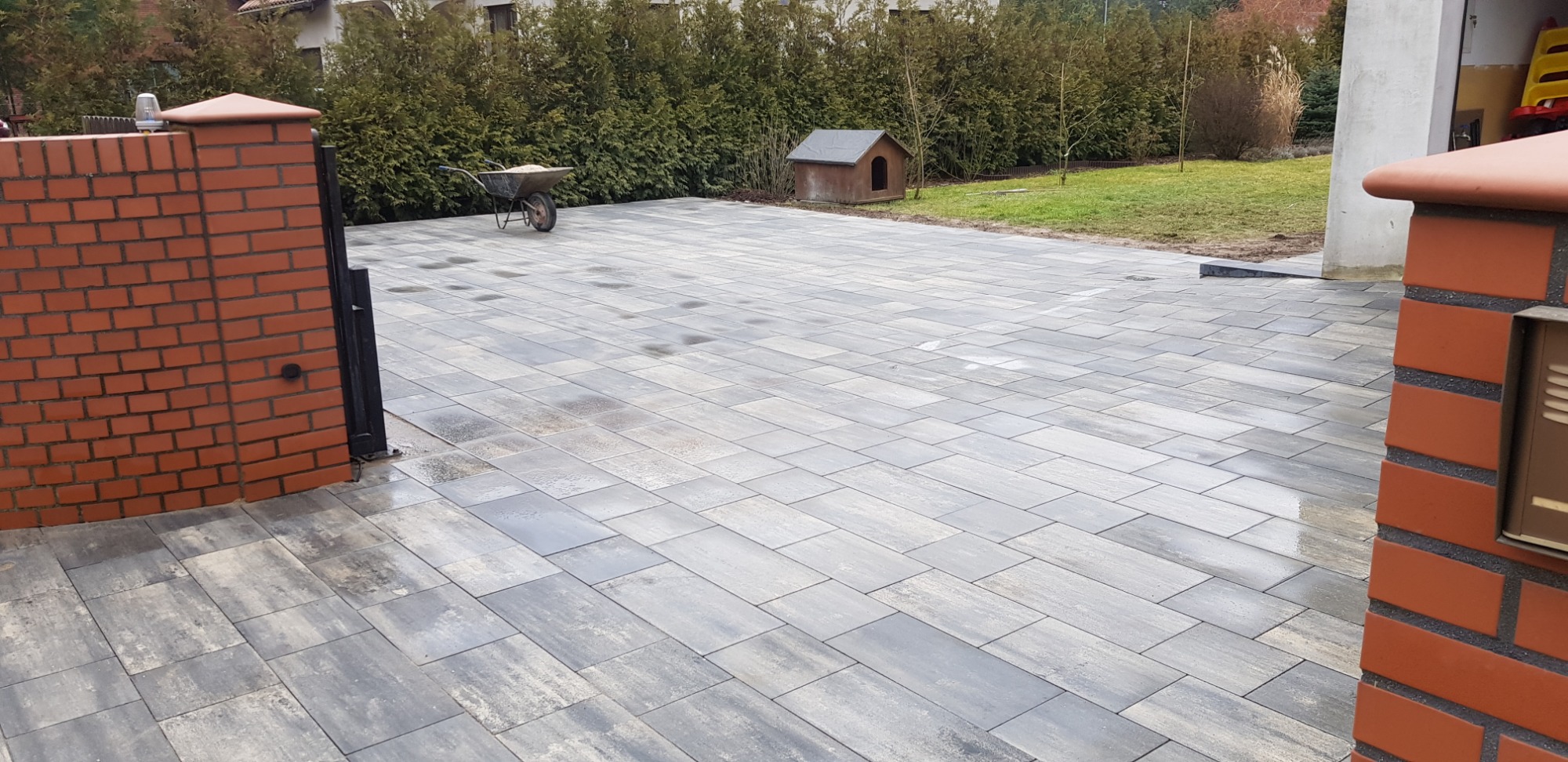 Nowo ułożona, wilgotna nawierzchnia z szarych prostokątnych płyt betonowych, z ceglanymi słupkami ogrodzenia i taczką w tle oraz budą dla psa, otoczona zielenią.