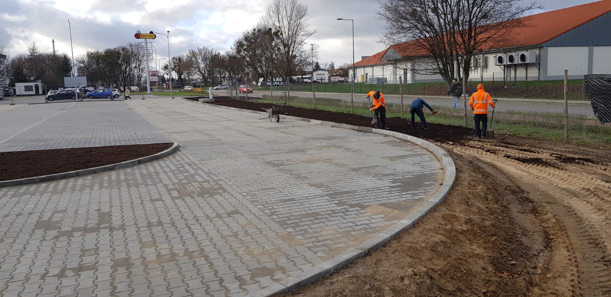 Układanie szarej kostki brukowej na parkingu sklepu, widoczni pracownicy w odblaskowych ubraniach wyrównujący ziemię pod nasadzenia wzdłuż obrzeża.