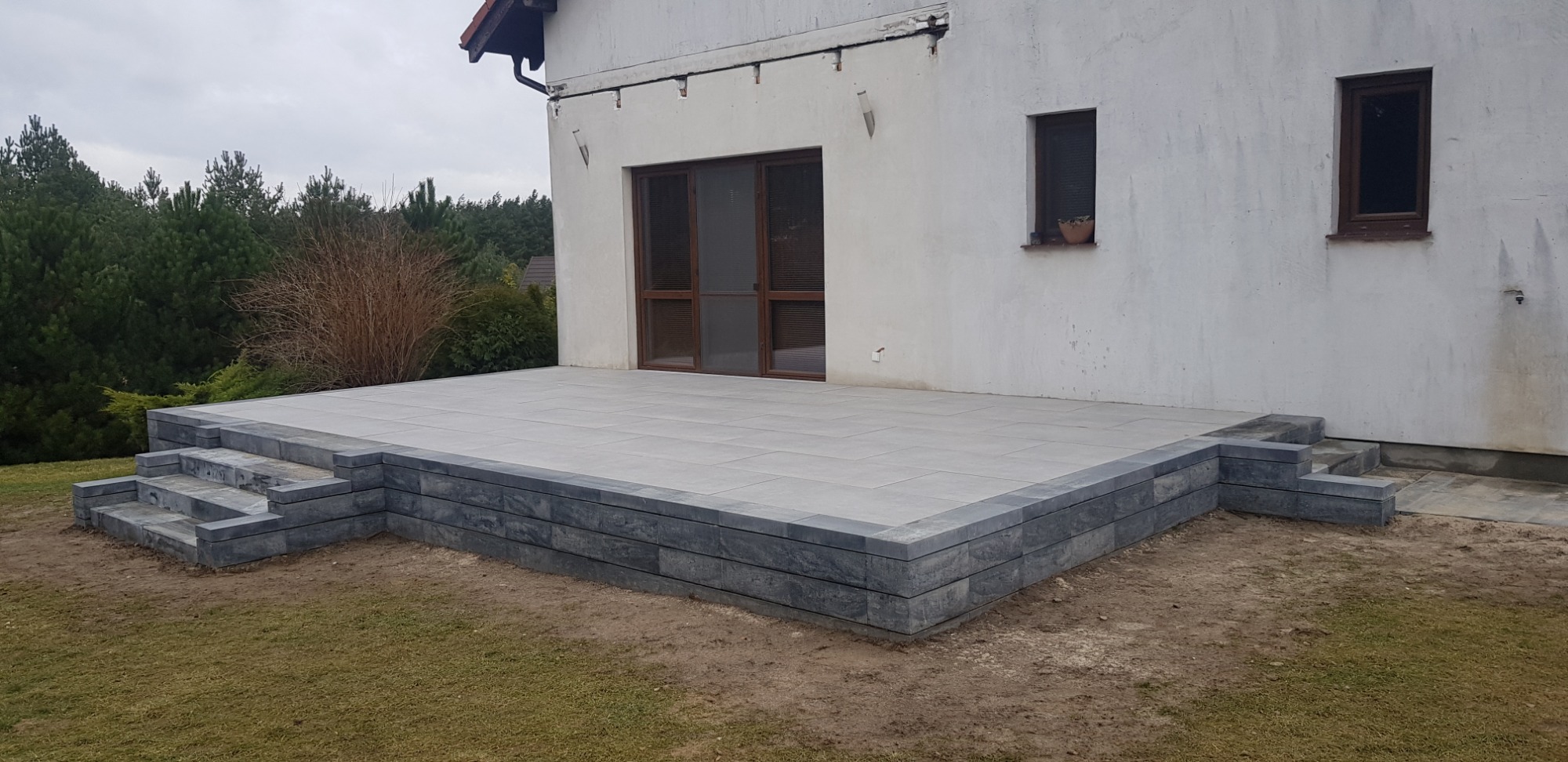 Szary taras z kostki brukowej, otoczony obrzeżem z ciemniejszej kostki, z niskimi schodami, przylegający do białego budynku z brązowymi oknami.