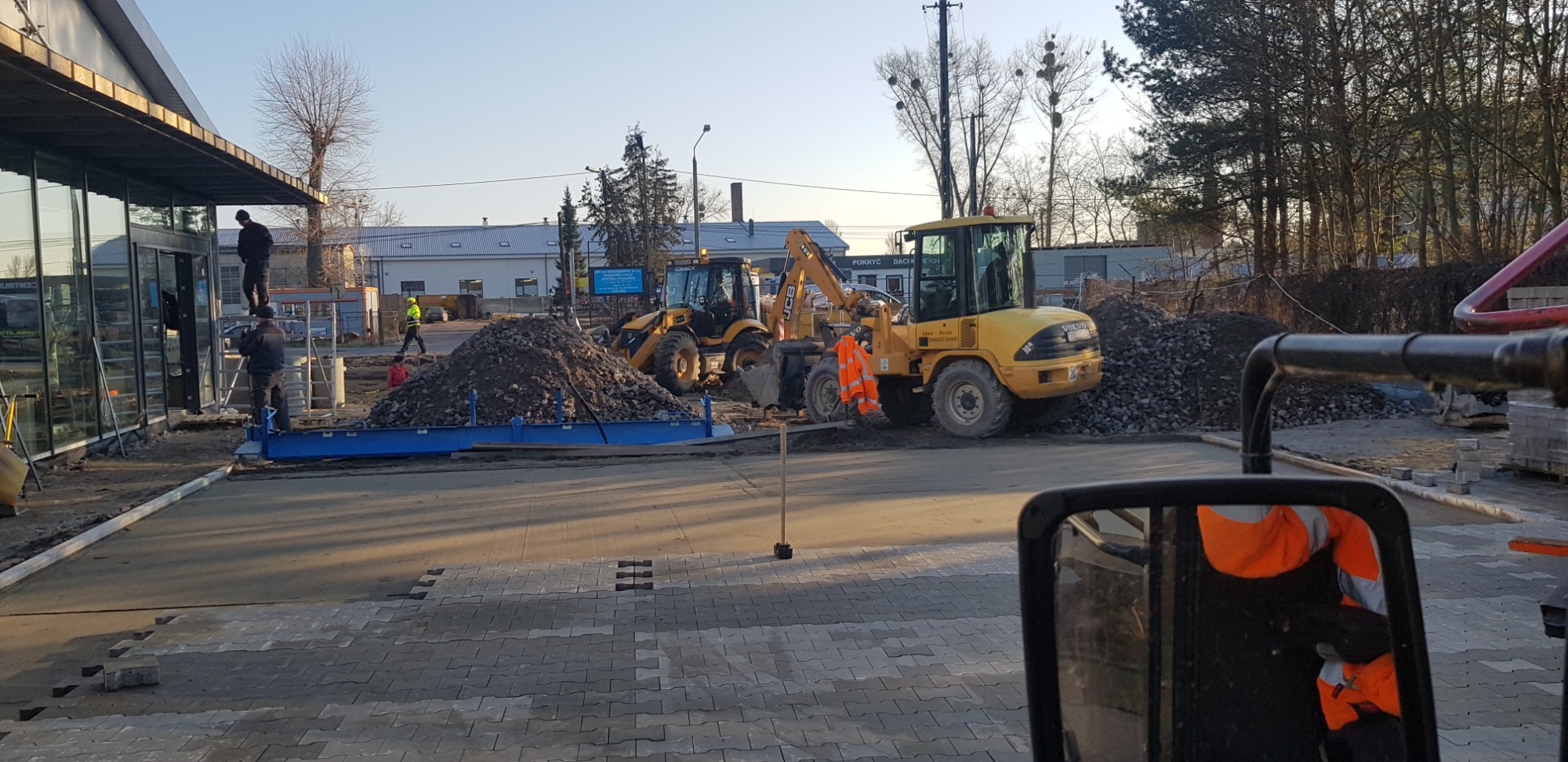 Układanie szarej kostki brukowej na placu przed budynkiem, z widocznymi maszynami budowlanymi i stertą gruzu, pracownicy w odblaskowych kamizelkach w tle, częściowo widoczne odbicie pracownika...