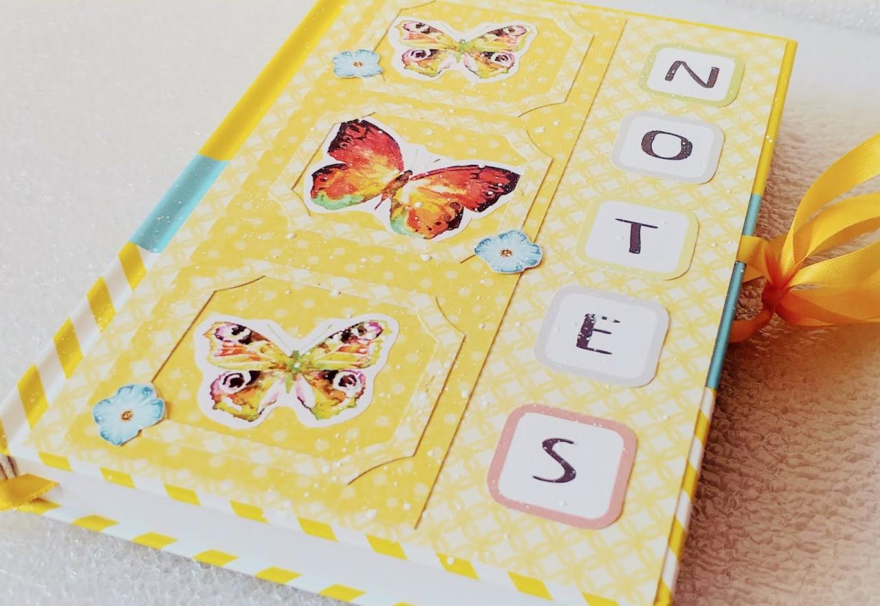 Żółty notes z motywem motyli i napisem 'NOTES', ozdobiony wstążką, leżący na białym tle.