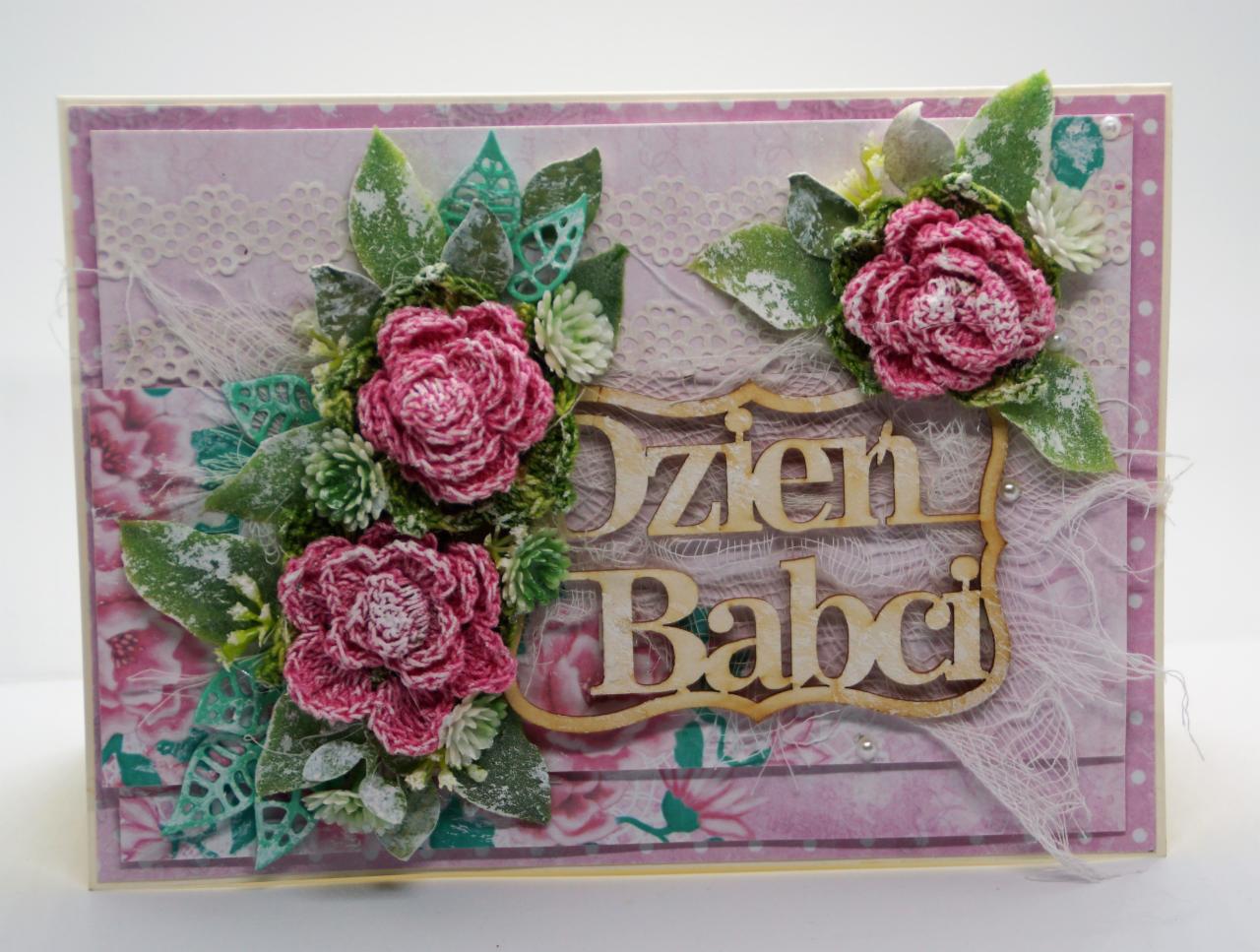 Ręcznie robiona kartka okolicznościowa z napisem 'Dzień Babci', ozdobiona trójwymiarowymi kwiatami z włóczki, koronką i filigranowymi wycinankami na różowym tle.