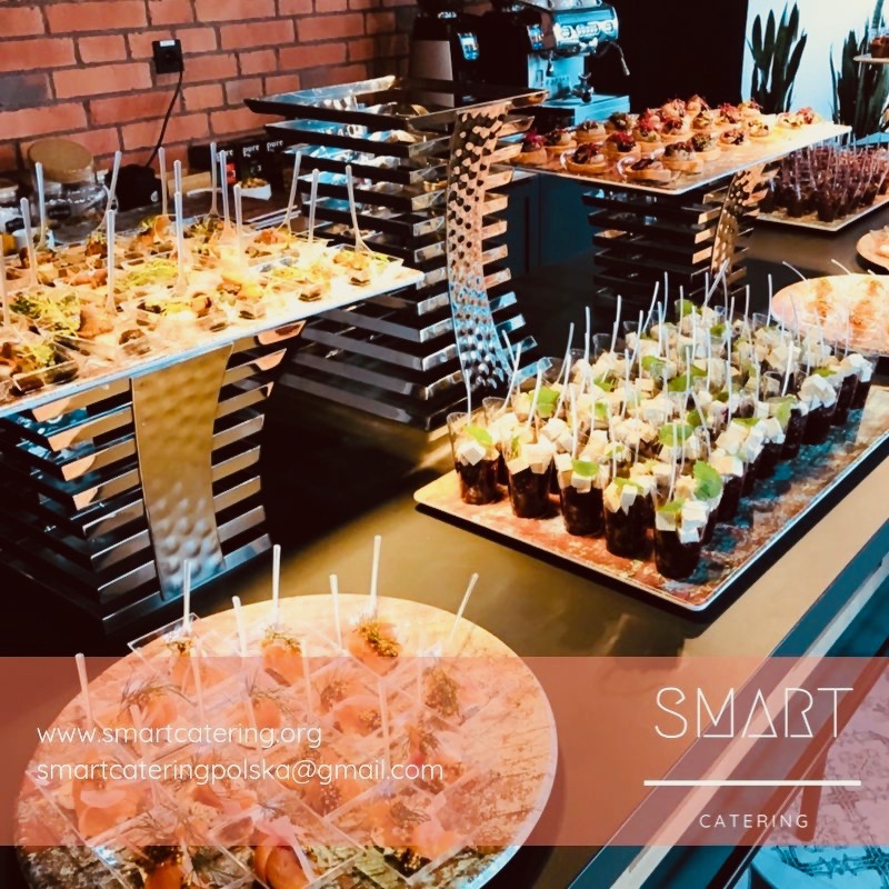 Elegancko zaaranżowany stół z różnorodnymi przekąskami na przyjęciu, w tle ceglana ściana i ekspres do kawy, widoczne logo firmy cateringowej w prawym dolnym rogu.