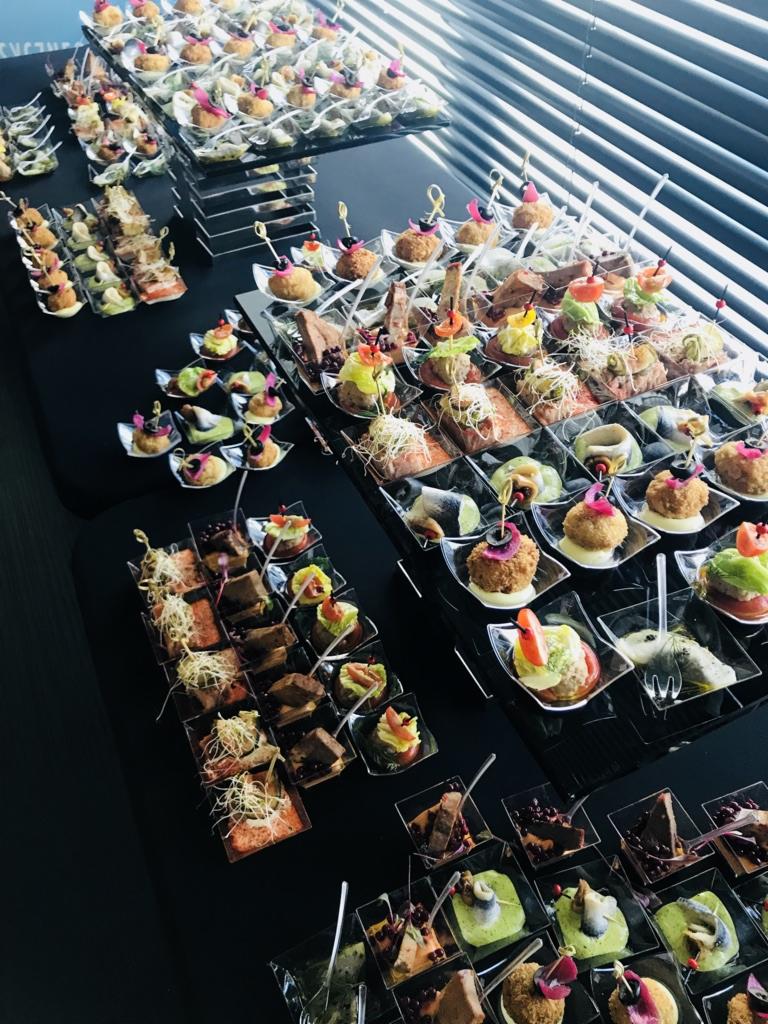 Elegancko zaaranżowany catering: różnorodne przekąski w przezroczystych, kwadratowych naczyniach, ułożone na czarnym stole z widocznym odbiciem, w tle żaluzje okienne.