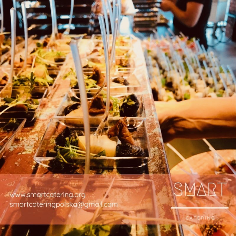 Rzędy przezroczystych pojemników z przekąskami na stole, widoczne sałatki, suszone śliwki, ser i inne dodatki, plastikowe widelczyki w każdym pojemniku, logo firmy w prawym dolnym rogu.