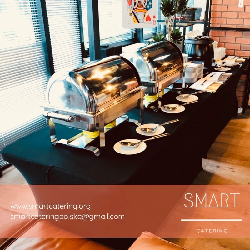 Stół cateringowy w Warszawie z dwoma podgrzewaczami do potraw, ułożonymi talerzami i sztućcami, widok z boku, w tle okno z żaluzjami i dekoracja z kartą królowej.