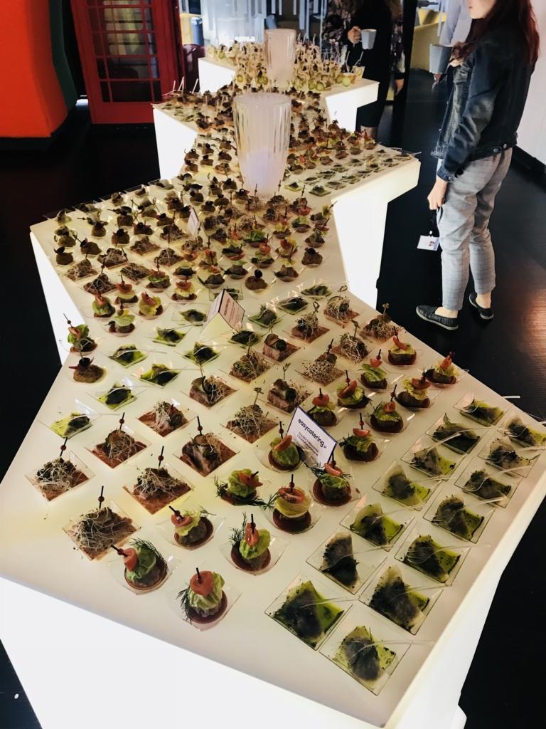 Starannie ułożone, różnorodne przekąski na białym, podświetlanym stole w kształcie gwiazdy, z widoczną czerwona budką telefoniczną w tle i osobami w otoczeniu.