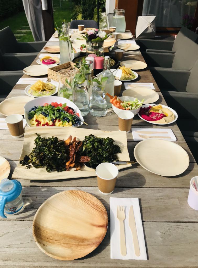 Długi stół z drewnianym blatem, zastawiony różnorodnymi daniami wegańskimi w naturalnych naczyniach, w tym sałatką owocową, pieczonymi warzywami korzeniowymi i chipsami z jarmużu, otoczony...