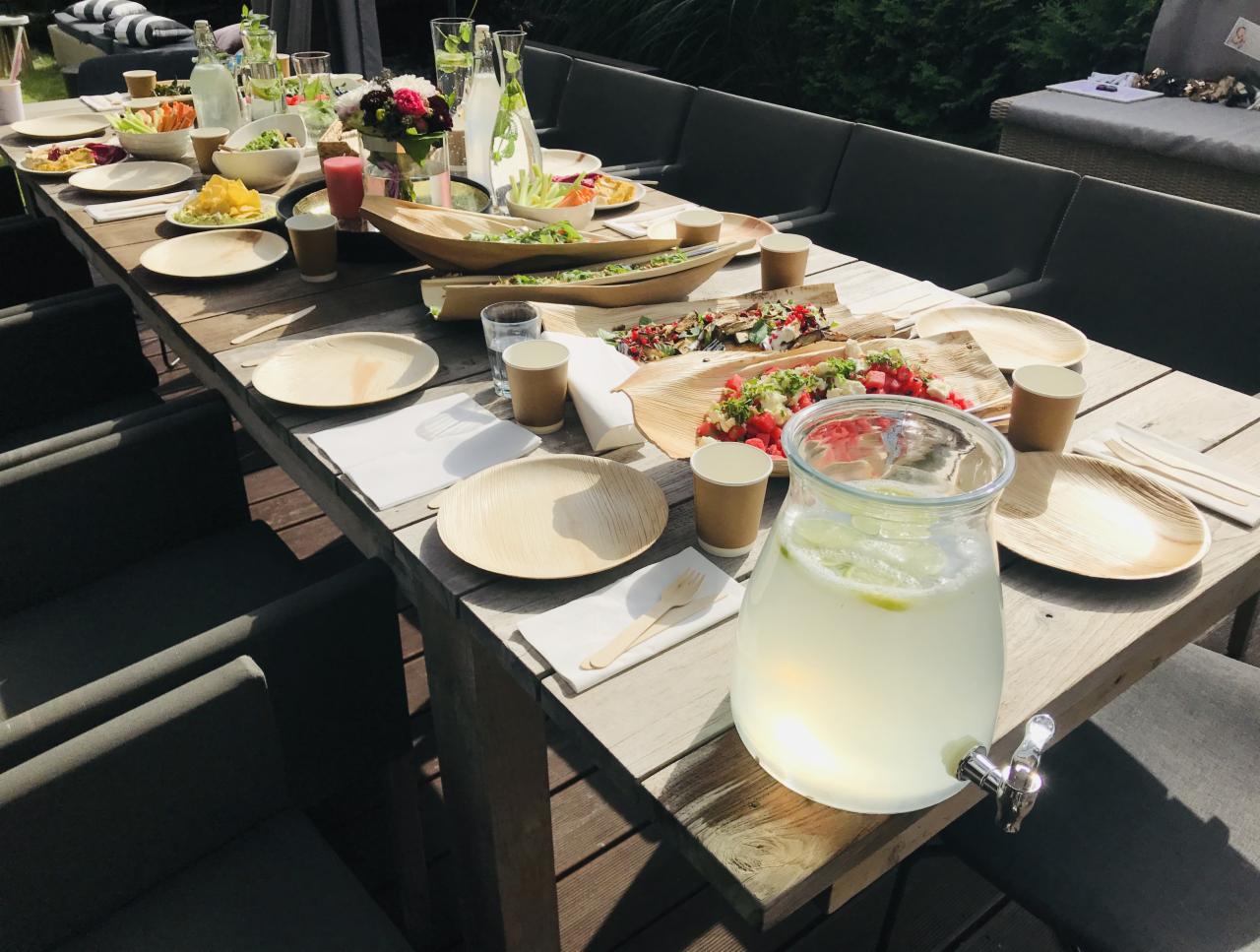 Stół na zewnątrz, nakryty ekologicznymi naczyniami, z sałatkami w drewnianych misach i dzbankiem lemoniady z kranikiem, przygotowany do przyjęcia.