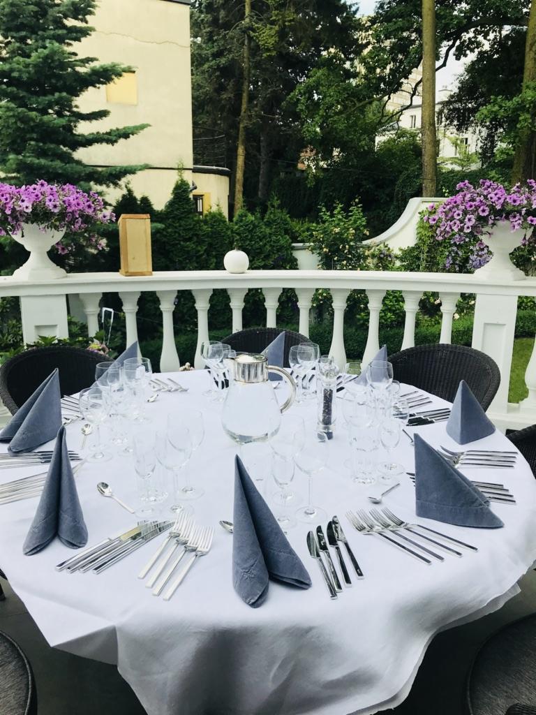 Nakryty okrągły stół na zewnątrz z szarą zastawą, srebrnymi sztućcami, szklankami i dzbankiem wody, otoczony zielenią i białymi balustradami z kwiatami.