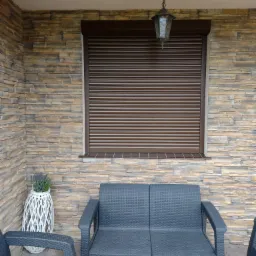Brązowa roleta zewnętrzna zamontowana w oknie na elewacji z kamienia dekoracyjnego w Gowarzewie, widok z zewnątrz na taras z meblami ogrodowymi.
