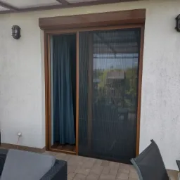 Zewnętrzna moskitiera plisowana w brązowej ramie na tarasie, widok z zewnątrz, szara sofa ogrodowa w kadrze.