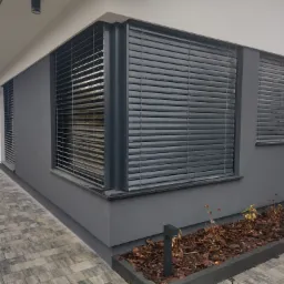 Elewacja domu z szarymi żaluzjami zewnętrznymi, widok z boku na narożne okno i chodnik z kostki brukowej.