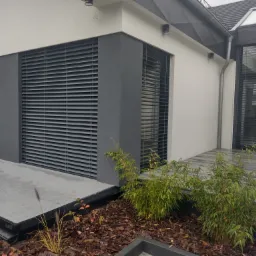 Szare poziome osłony okienne zewnętrzne na elewacji nowoczesnego domu z szarą kostką i rabatą z korą i zieloną roślinnością.