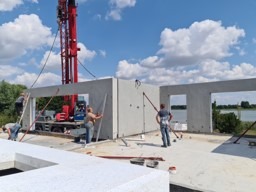 Wznoszenie ścian z prefabrykatów betonowych na placu budowy z widokiem na jezioro, robotnicy w trakcie montażu, dźwig w tle.