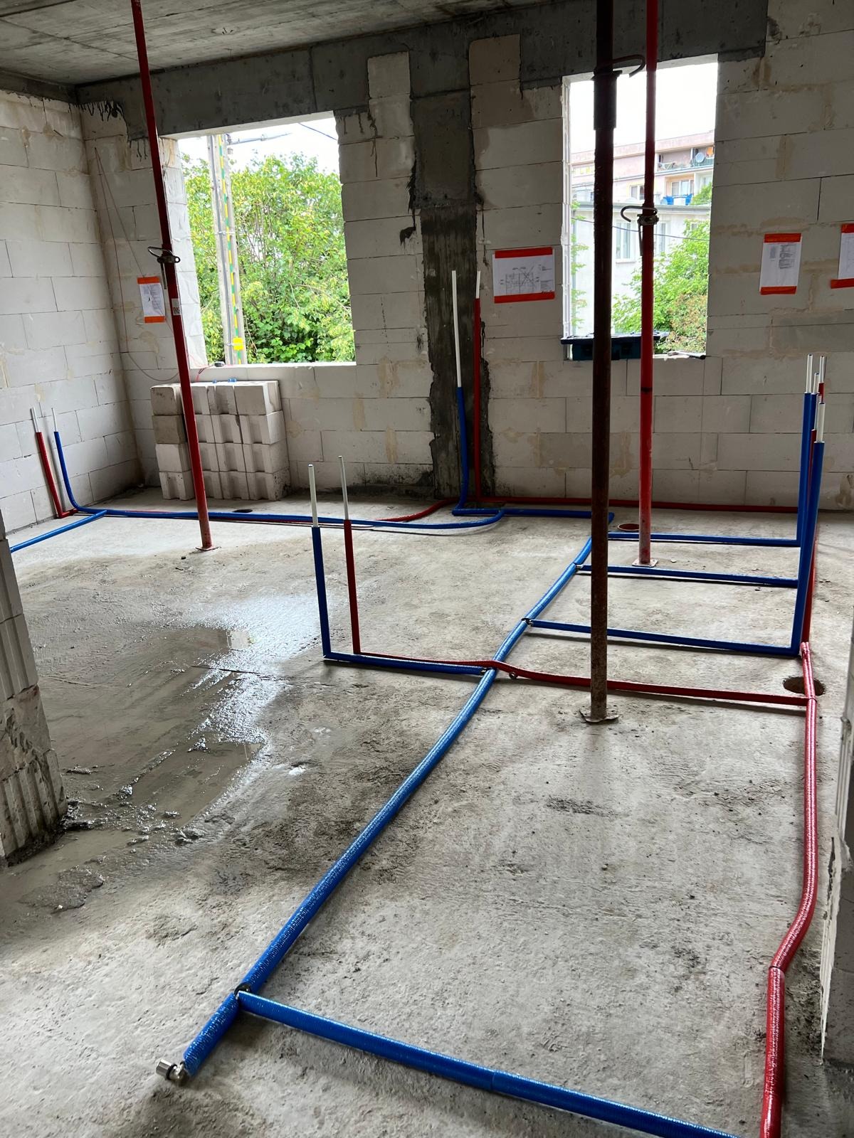 Wnętrze w stanie surowym z widocznymi instalacjami wodnymi: niebieskie i czerwone rury prowadzone po podłodze, pionowe podpory budowlane oraz ściany z białego bloczka.