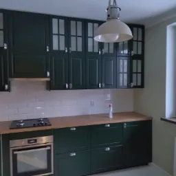 Nowoczesna kuchnia z ciemnozielonymi szafkami, białymi płytkami na ścianie i drewnianym blatem, oświetlona wiszącą lampą w stylu industrialnym, z widokiem na zewnątrz przez okno.