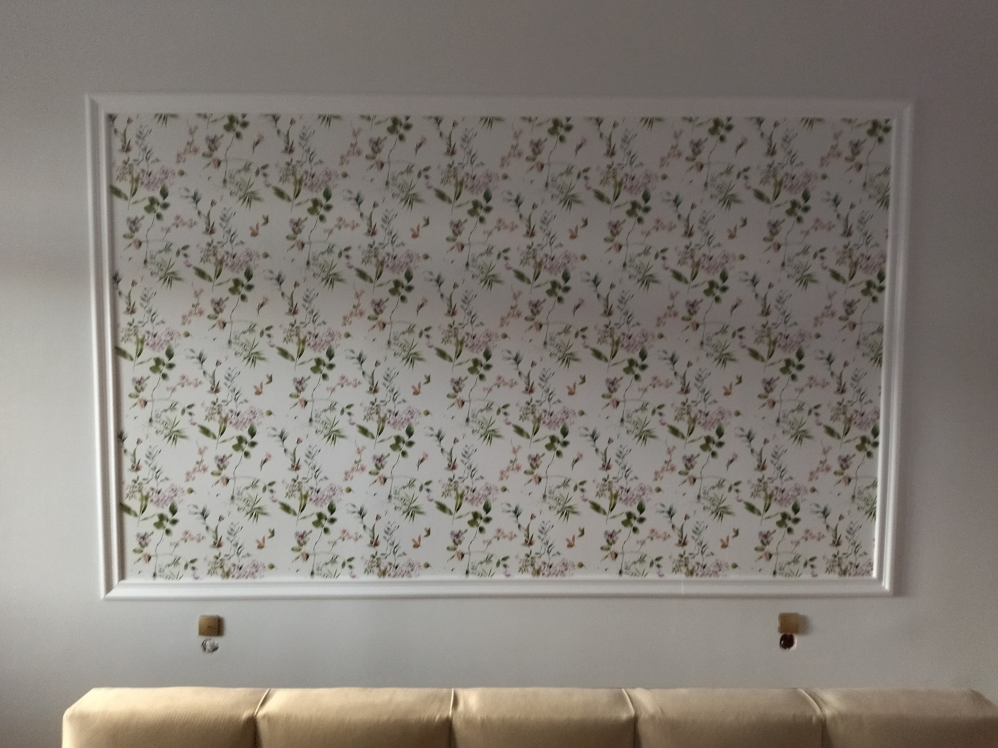 Wzorzysta tapeta w ramie z białego sztukateria, widoczny fragment sofy w neutralnym odcieniu.