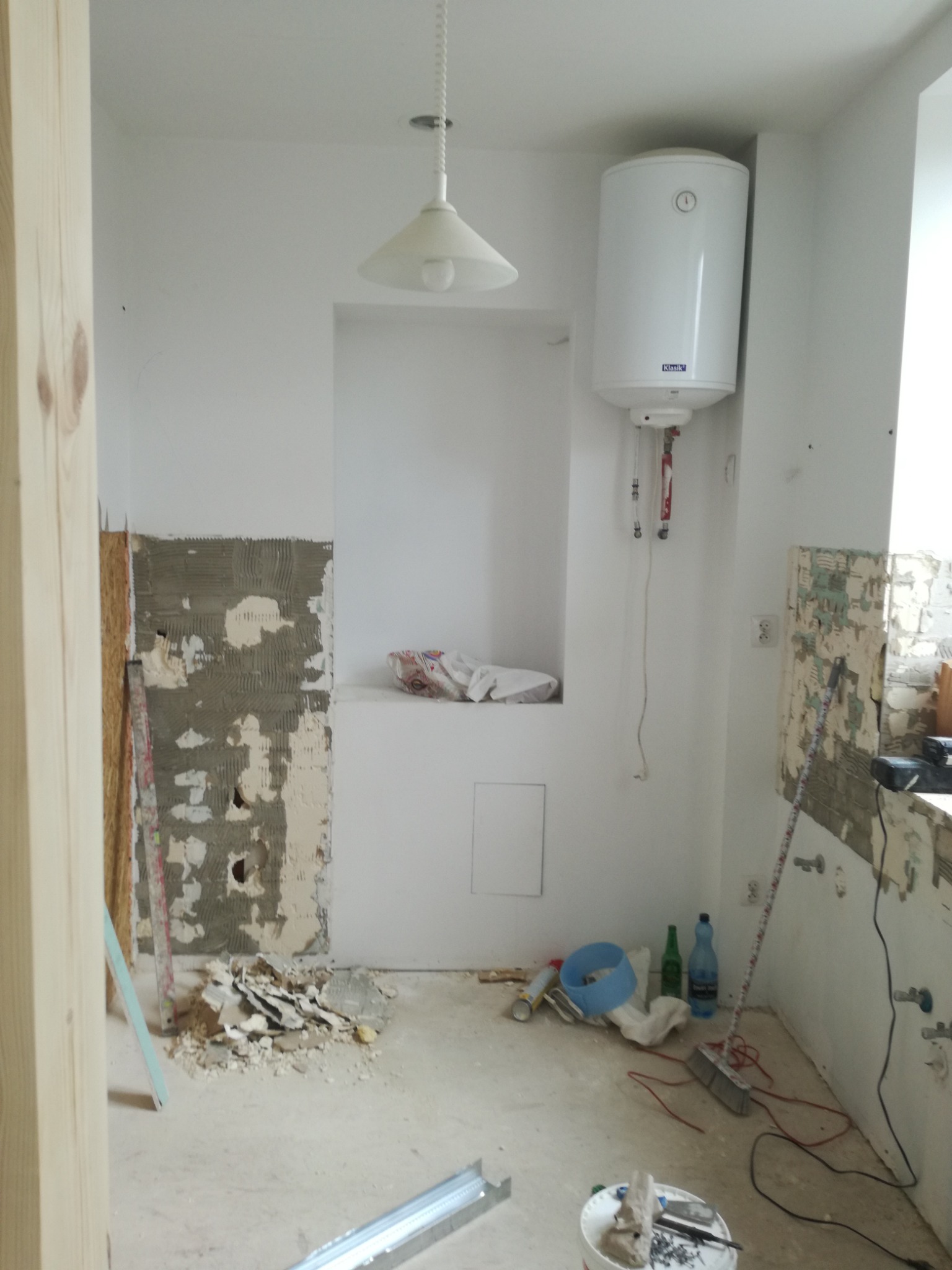 Wnętrze łazienki w trakcie demontażu starych płytek, widoczny bojler, resztki gruzu i narzędzia na podłodze, przygotowania do dalszych prac.