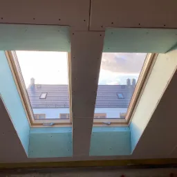 Dwa okna dachowe z widokiem na budynki we Wrocławiu, obudowane surową płytą kartonowo-gipsową w trakcie montażu, widoczne łączenia i wkręty.