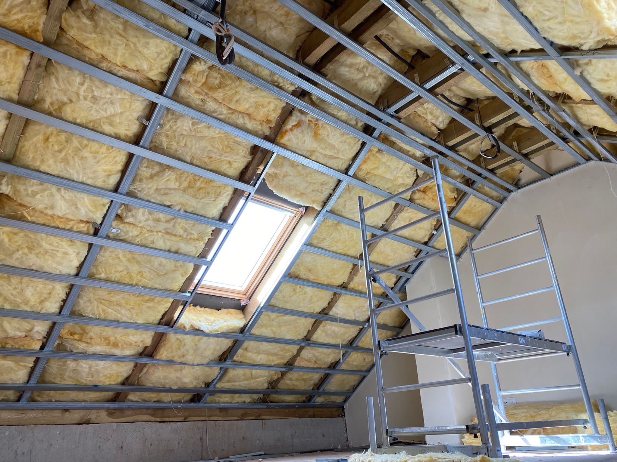 Ocieplenie poddasza wełną mineralną, widoczna metalowa konstrukcja nośna i okno dachowe, drabina aluminiowa w tle.