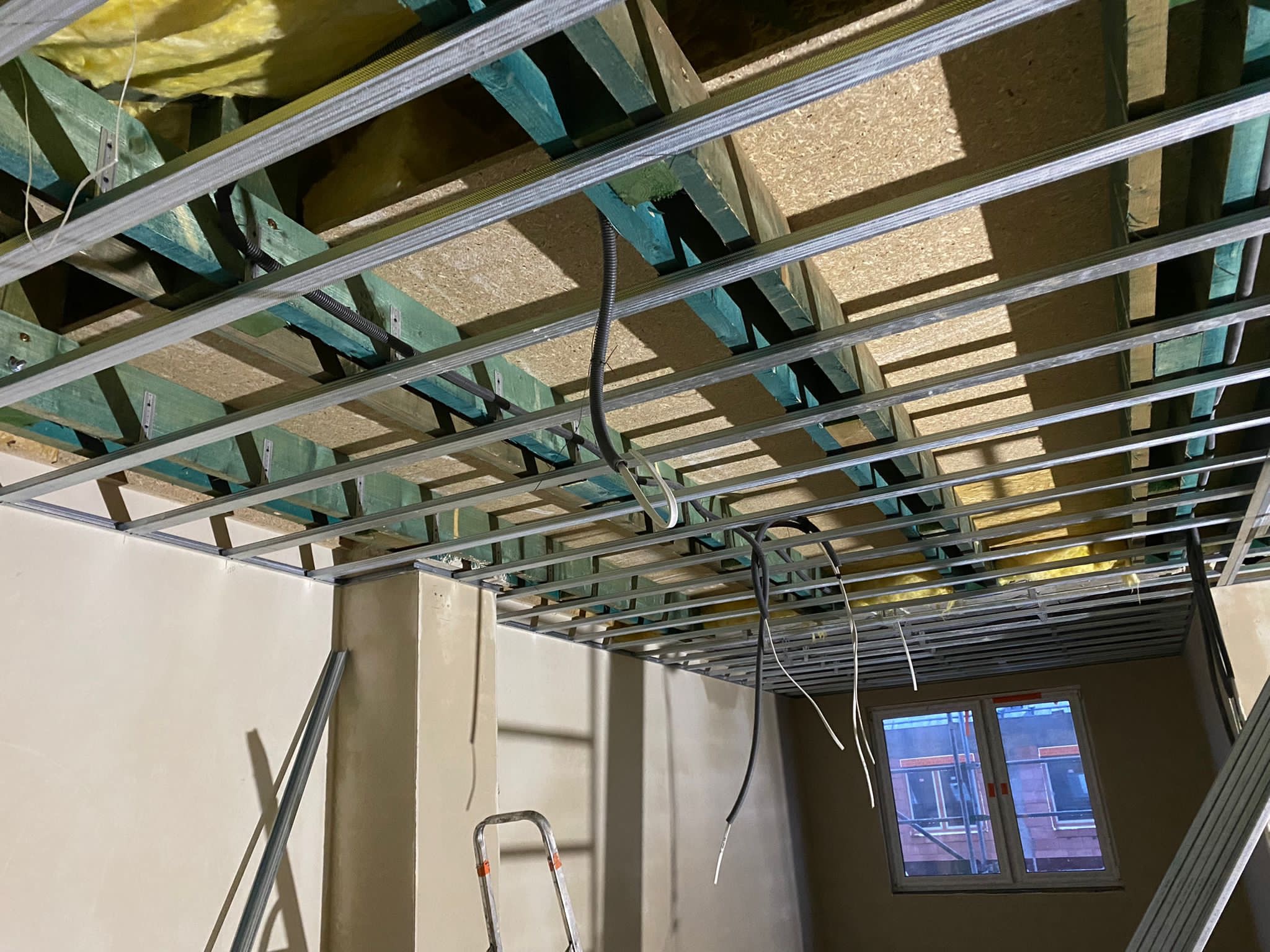 Montaż metalowej konstrukcji sufitu podwieszanego z widoczną izolacją, drewnianymi belkami i przewodami elektrycznymi w pomieszczeniu z oknem.