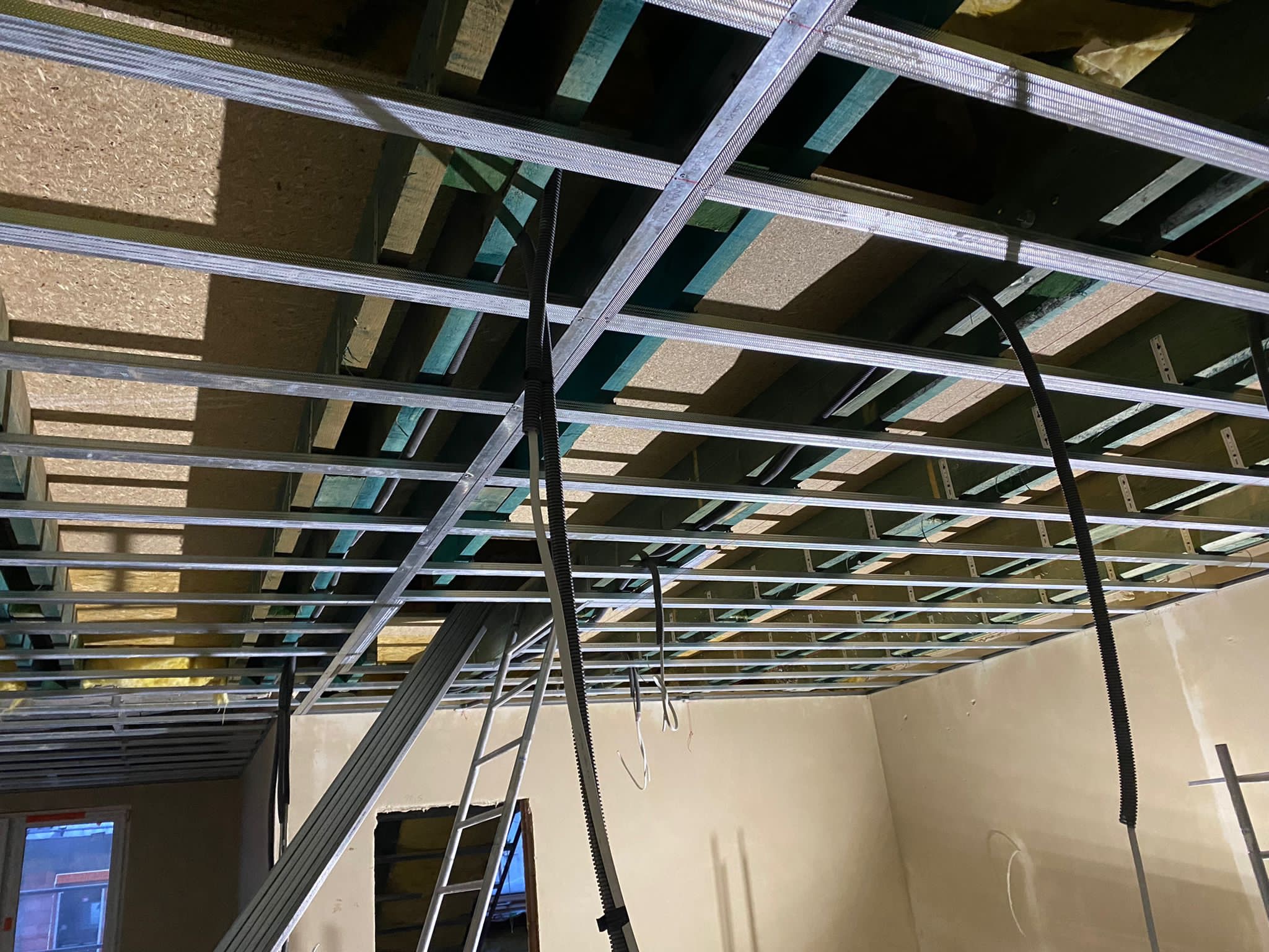 Metalowa konstrukcja sufitu podwieszanego z widoczną izolacją, czarne kable zwisające z góry, drabina oparta o ścianę w remontowanym pomieszczeniu.