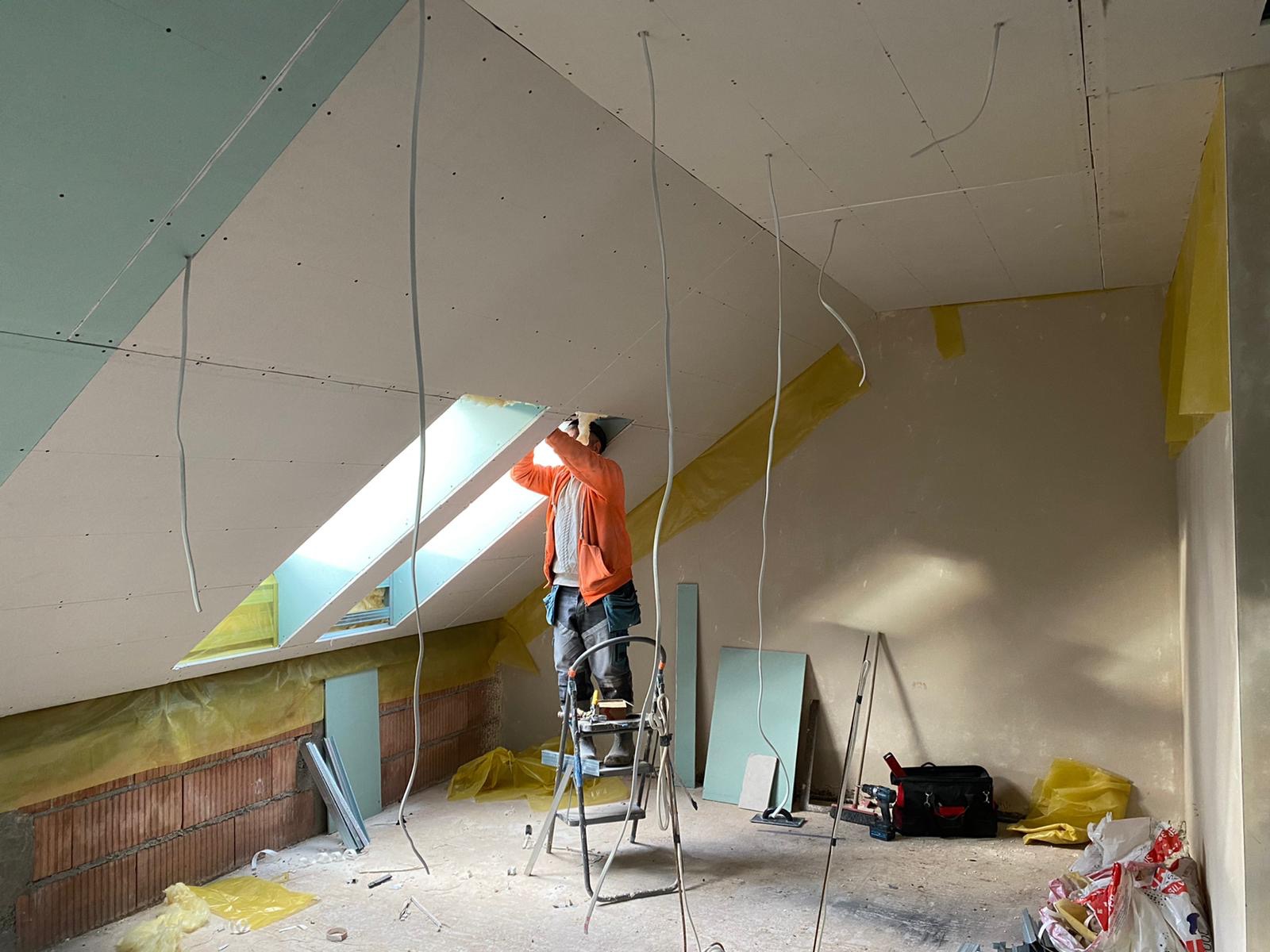 Montaż płyty gipsowo-kartonowej przy oknie dachowym w pokoju na poddaszu, widoczny pracownik na drabinie, instalacja elektryczna wystająca z sufitu, oraz narzędzia i materiały budowlane.