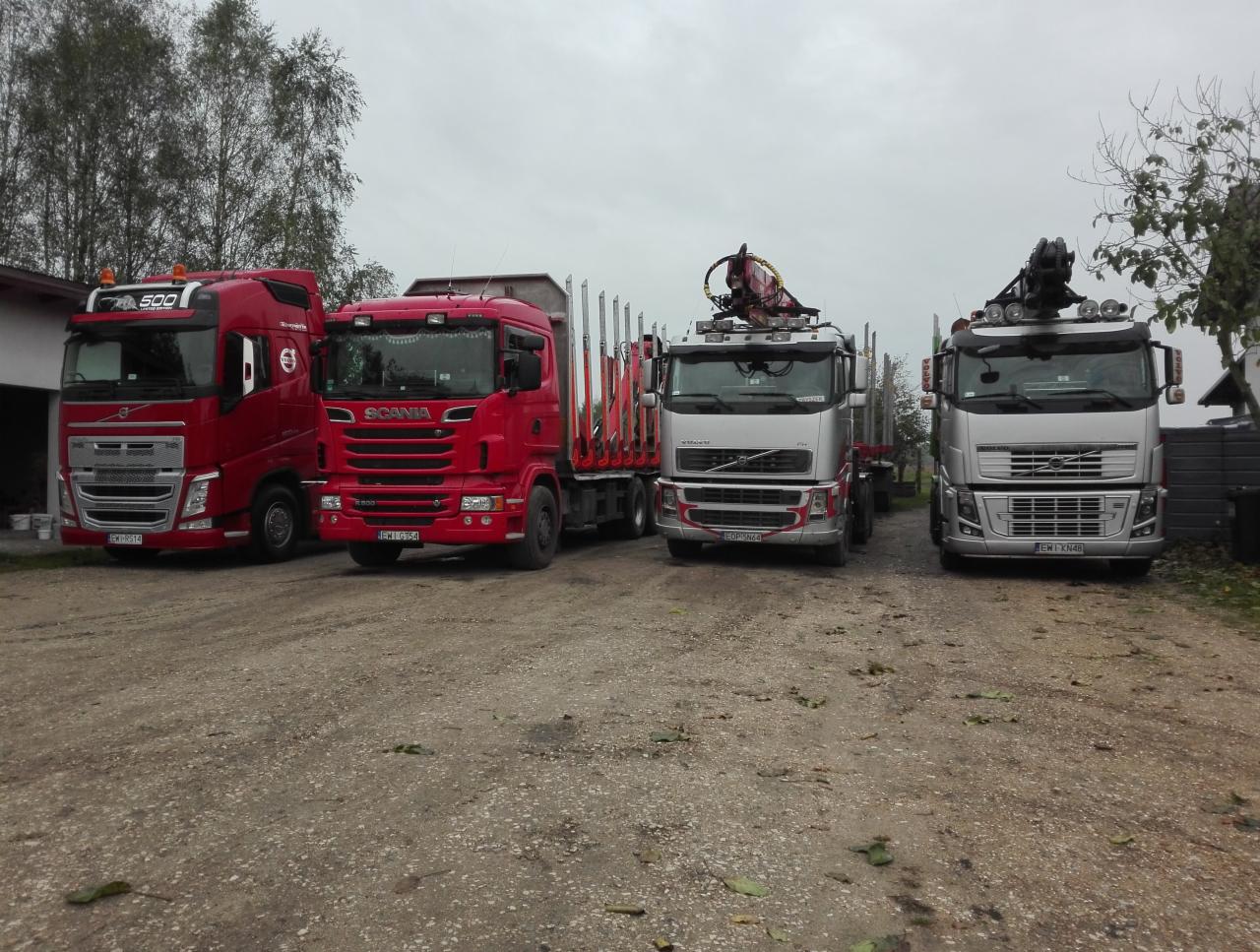 Cztery ciężarówki: czerwony Volvo FH 500, czerwona Scania z przyczepą do transportu drewna, srebrny Volvo z dźwigiem HDS do załadunku drewna i srebrny Volvo z dźwigiem HDS na szarym, żwirowym placu.