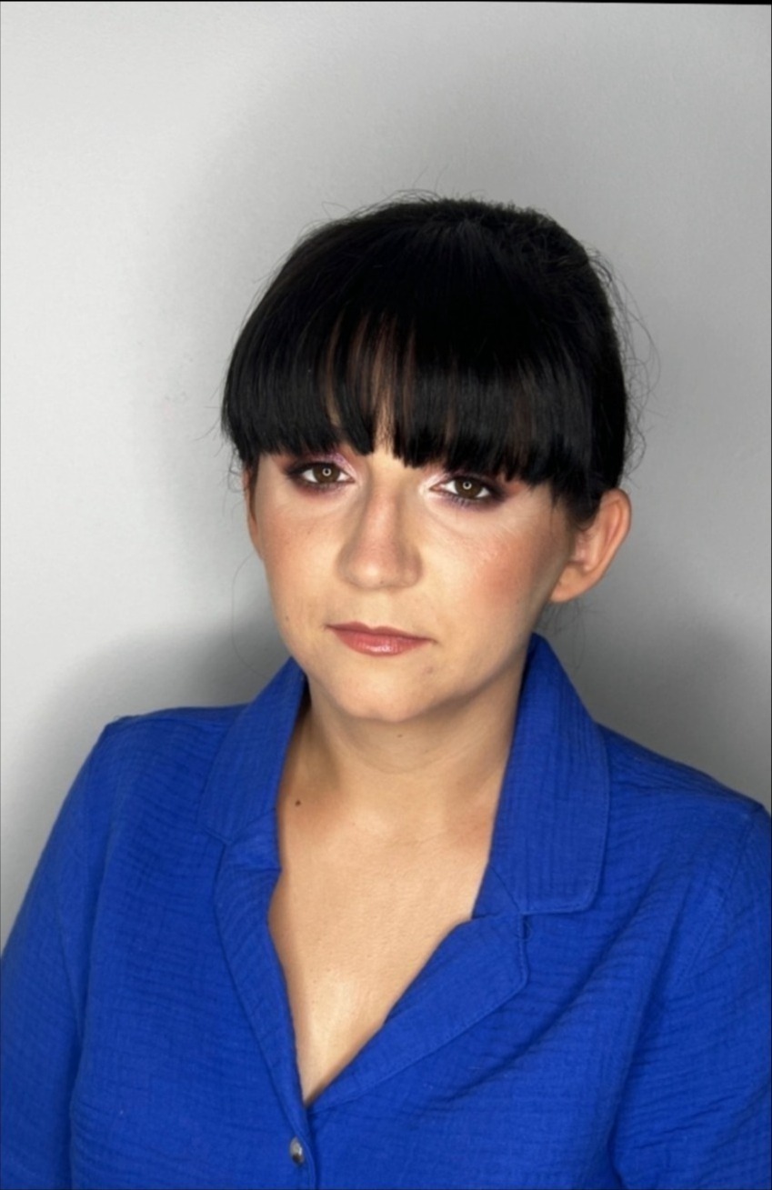 Portret kobiety z wyrazistym makijażem oczu w odcieniach brązu i różu, podkreślonymi policzkami i delikatnym błyszczykiem, ubrana w niebieską koszulę.