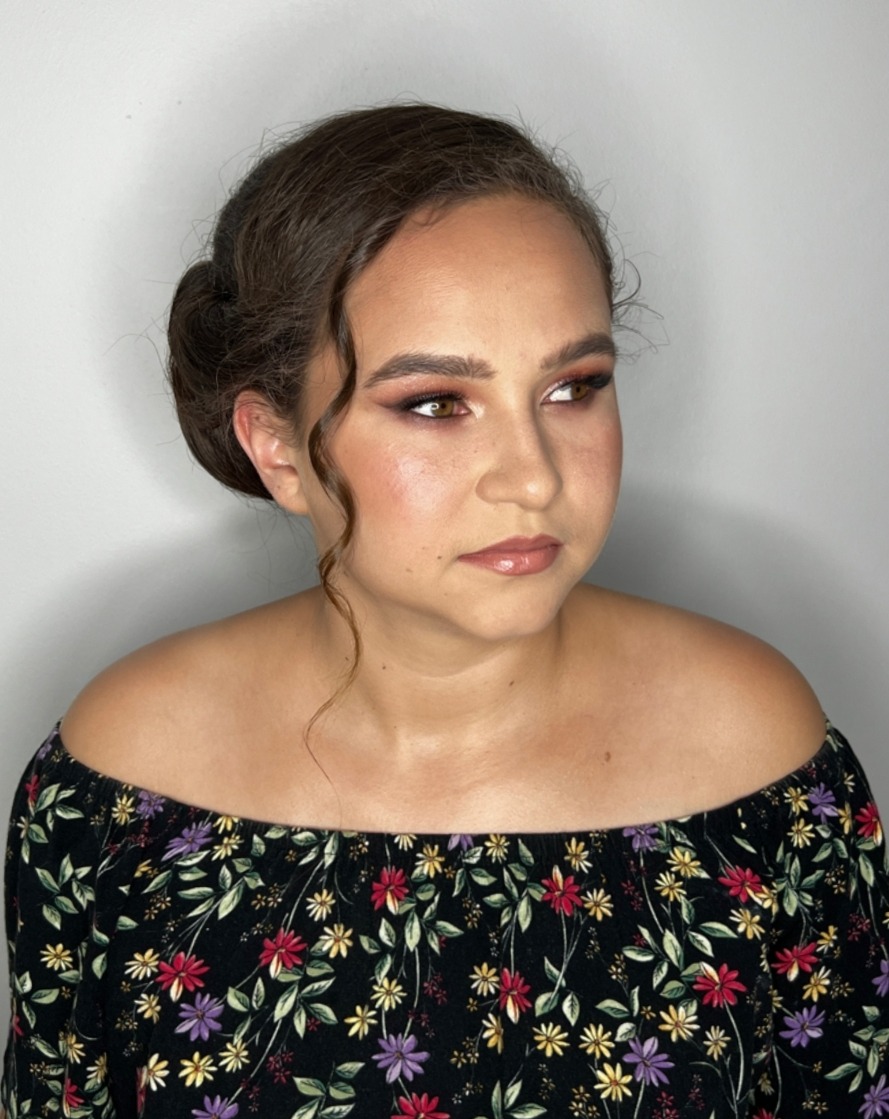 Portret kobiety z delikatnym makijażem oczu i ust, upiętymi włosami, ubrana w bluzkę w kwiaty, spoglądającej w bok na jasnym tle.