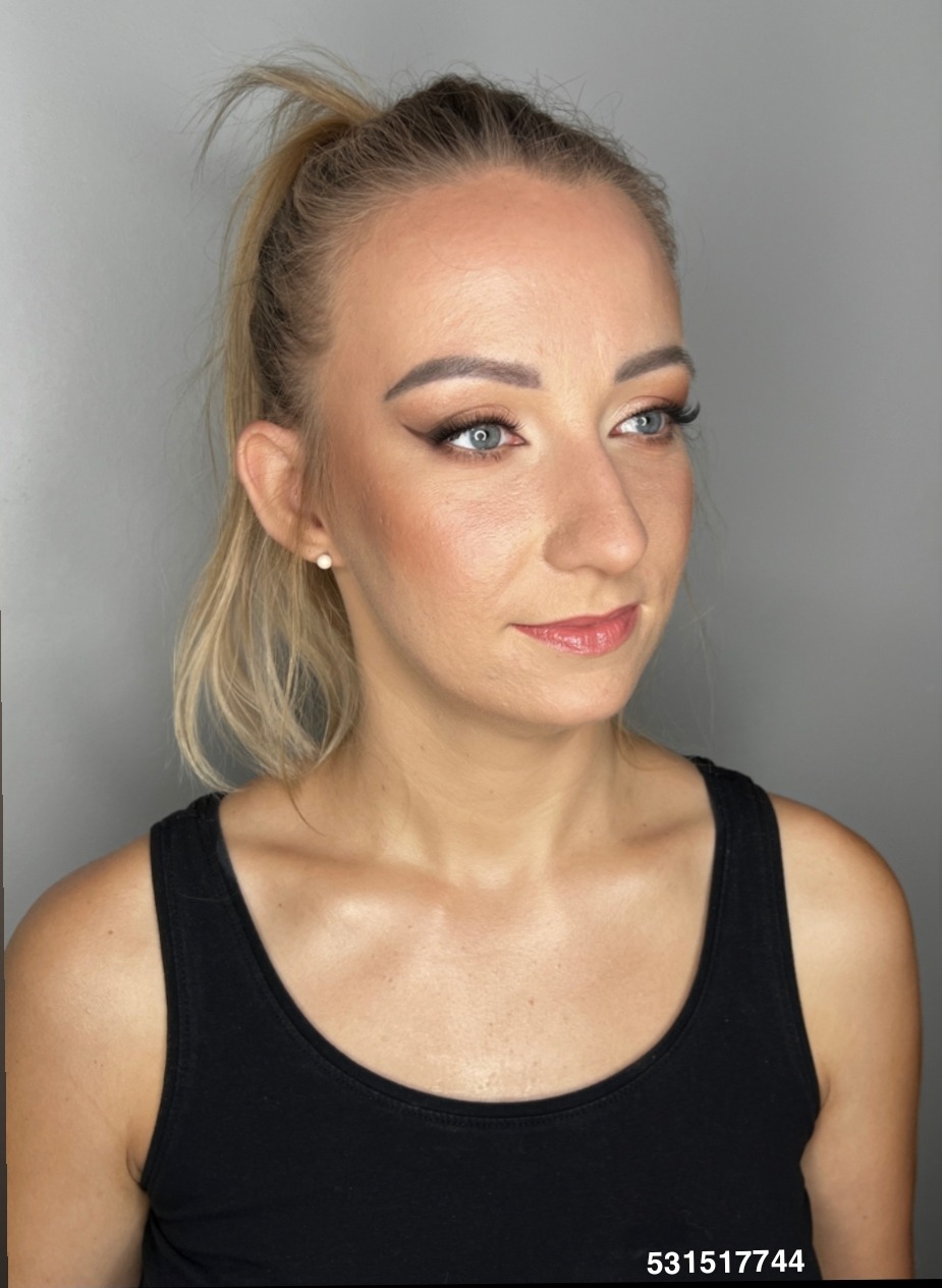 Portret kobiety z delikatnym makijażem, podkreślone oczy eyelinerem, włosy spięte w kucyk, subtelny rumieniec i błyszczyk. Jasna karnacja, szare tło.