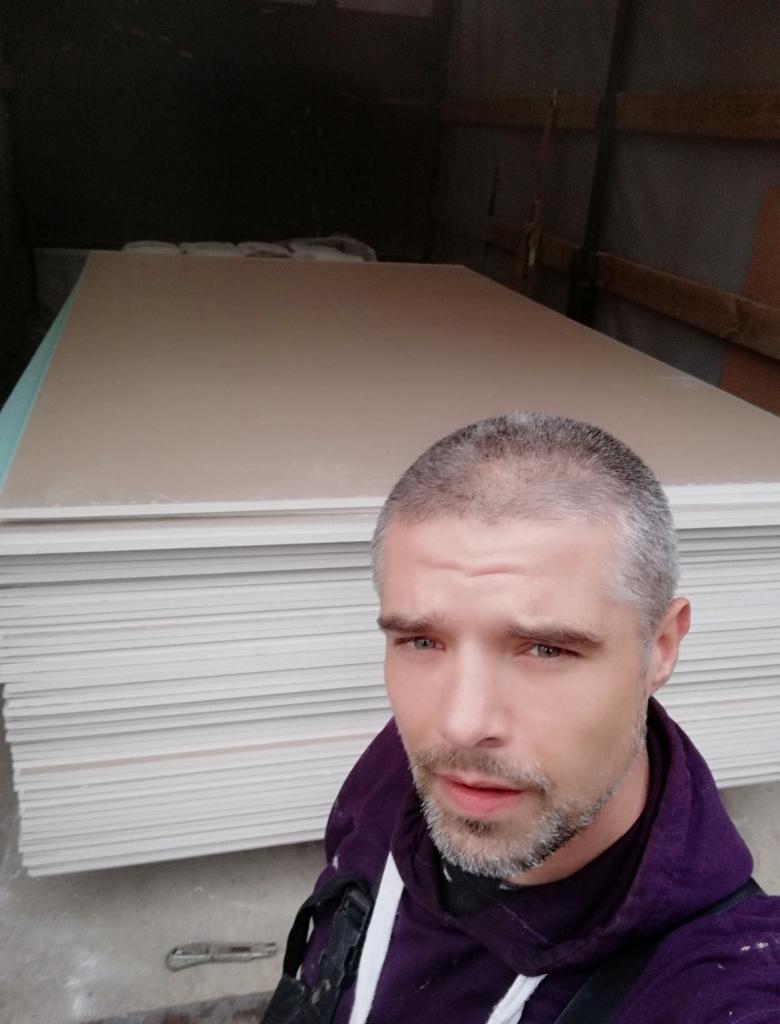 Mężczyzna robi selfie na tle stosu płyt karton-gips w szarym pomieszczeniu. Widoczny nóż do cięcia płyt. Ubrany w fioletową bluzę.