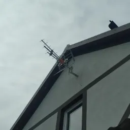 Antena telewizyjna zamontowana na ścianie szczytowej domu jednorodzinnego z widocznym oknem i ptakiem siedzącym na dachu, sfotografowana z dołu przy pochmurnym niebie.