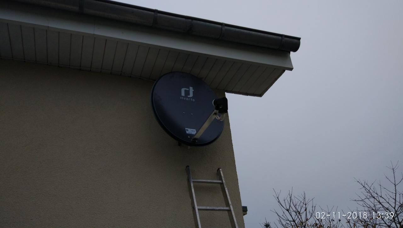 Czarna antena satelitarna marki Inverto zamontowana na elewacji budynku, z przystawioną aluminiową drabiną, widoczna data i godzina na zdjęciu.