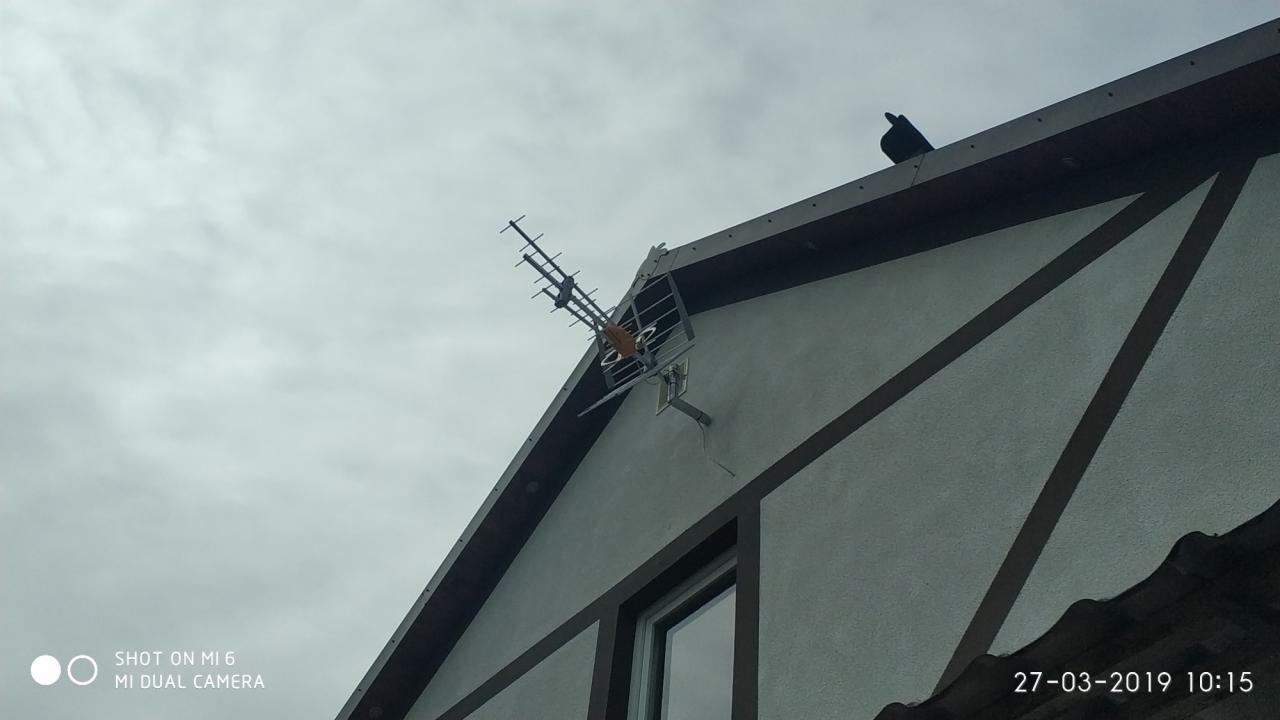 Antena telewizyjna zamontowana na ścianie szczytowej domu jednorodzinnego z widocznym oknem i ptakiem siedzącym na dachu, sfotografowana z dołu przy pochmurnym niebie.