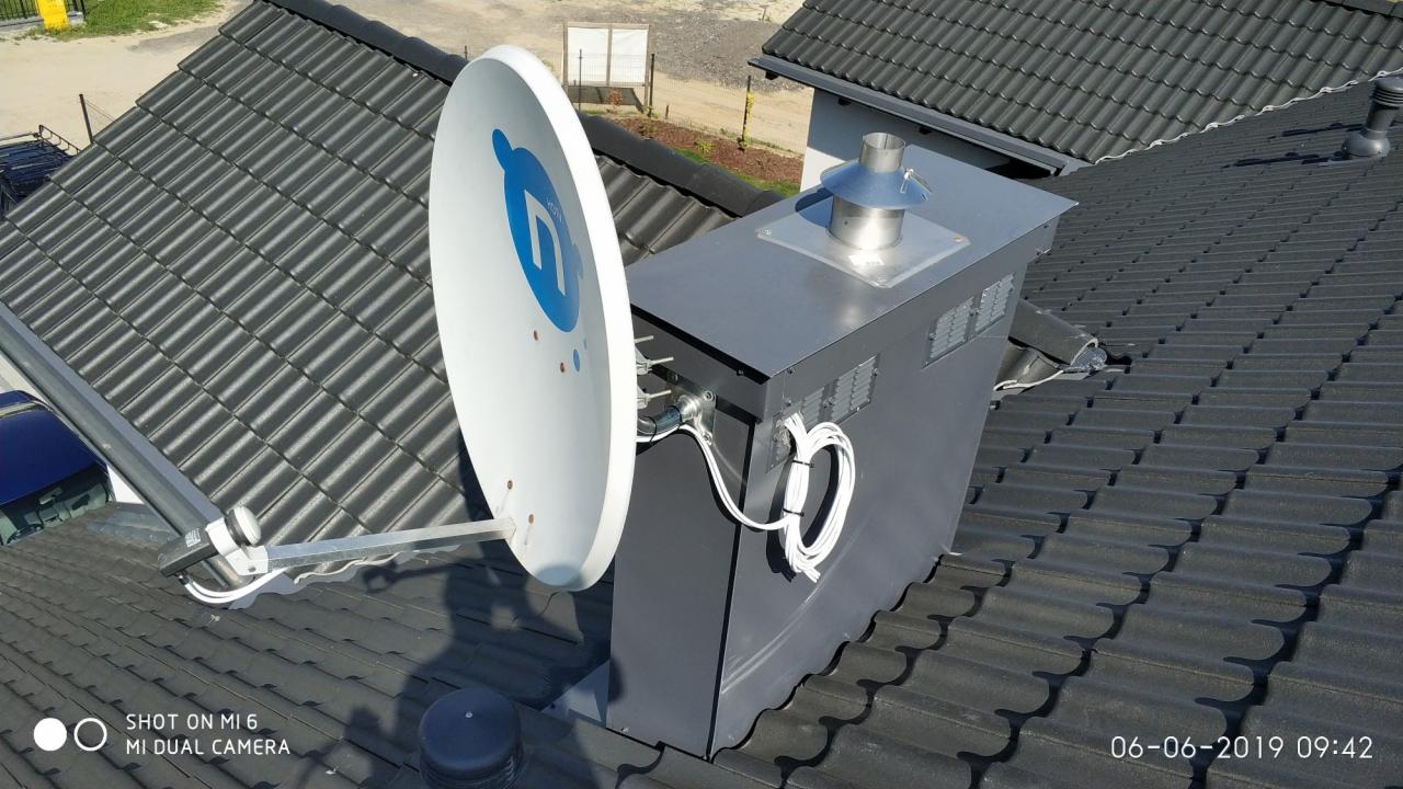 Antena satelitarna z niebieskim logo zamontowana na dachu z dachówki ceramicznej, obok komina z metalową obudową i widocznymi kablami.