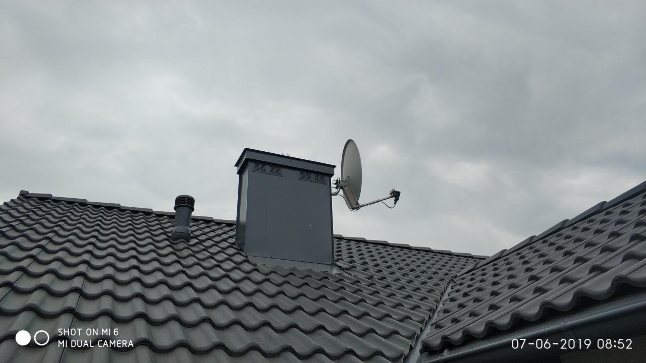 Antena satelitarna zamontowana na kominie wentylacyjnym na dachu z ciemnej dachówki ceramicznej, widok z dołu pod pochmurnym niebem.