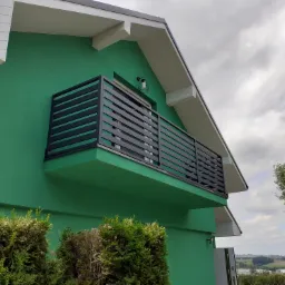 Nowoczesny balkon z czarną, poziomą balustradą na tle zielonej ściany domu i pochmurnego nieba.