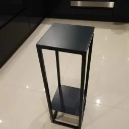 Czarny, metalowy kwietnik w minimalistycznym stylu, z dwoma poziomami, stojący na jasnych, błyszczących płytkach w kuchni z czarnymi szafkami i piekarnikiem w tle.