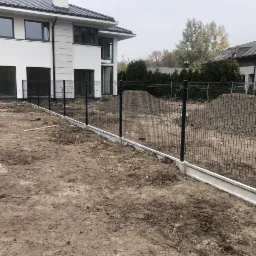 Montaż ogrodzenia panelowego na betonowej podmurówce przed nowoczesnym domem z białymi ścianami i ciemną dachówką, widoczne kopce ziemi po pracach ziemnych.