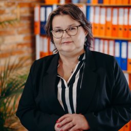 EUROMAX.PL SPÓŁKA Z OGRANICZONĄ ODPOWIEDZIALNOŚCIĄ SPÓŁKA KOMANDYTOWA - Portret kobiety w okularach i czarnym żakiecie na tle biura z regałem pełnym segregatorów w Gdańsku. Widoczne czerwone paznokcie i delikatna biżuteria.